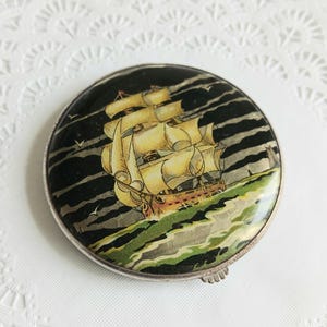 Peut inclure: Un compact vintage argenté avec une image colorée d'un navire naviguant sur une mer bleu foncé avec des nuages blancs. Le navire a des voiles blanches et une coque brune.