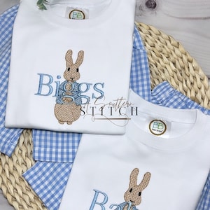 May include: Two white t-shirts with blue embroidered Peter Rabbit designs. The shirts are personalized with the names "Biggs" and "Baker".