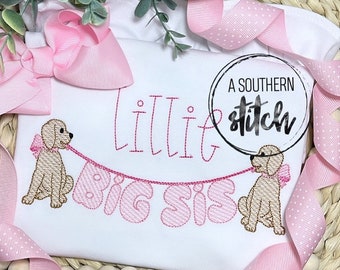 Puede incluir: Mono blanco con bordado rosa que dice "Lillie Big Sis" con dos perros marrones que sostienen pancartas rosas. El mono está rodeado de cinta rosa.