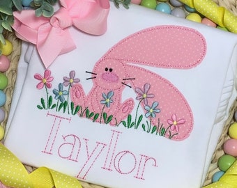 May include: A white onesie with a pink bunny embroidered on it. The bunny is surrounded by flowers and the name "Taylor" is embroidered below it.