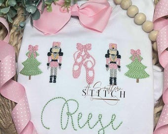 Könnte beinhalten: Weißer Strampler mit rosa Schleife, grünen gestickten Weihnachtsbäumen, einem Paar rosa Ballettschuhen und zwei Nussknackerfiguren. Der Strampler ist mit dem Namen "Reese" in grüner Stickerei personalisiert.