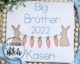 May include: A white babygrow with blue embroidered text that reads "Big Brother 2022" and a name "Kacen". Two brown bunnies are embroidered on either side of the text with a string of carrots hanging between them.