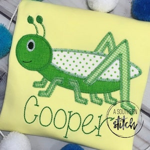 Puede incluir: Un pelele amarillo con una langosta bordada en verde y el nombre "Cooper" cosido en hilo verde. El pelele tiene un pequeño círculo blanco con el texto "A Southern Stitch" en negro.