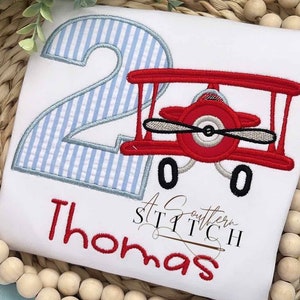 May include: A white onesie with a blue and white striped number two, a red aeroplane with a grey propeller, and the name "Thomas" embroidered in red. The text "A Southern Stitch" is also embroidered on the onesie.