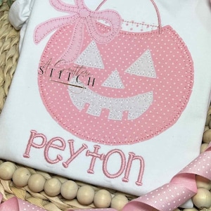 May include: A pink and white pumpkin with a bow and the text "A Cutters Stitch" and "peyton" embroidered on a white shirt.
