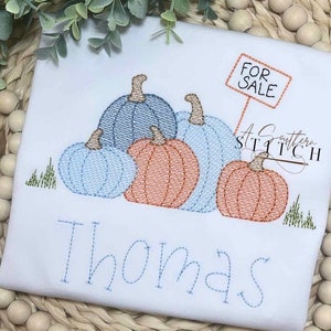 Peut inclure: Une grenouillère blanche avec un motif brodé de citrouilles et un panneau indiquant "À vendre". La grenouillère porte également le nom "Thomas" brodé en bas.