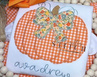 May include: A white onesie with an orange and white gingham pumpkin with a floral bow. The onesie has the name "ava drew" embroidered in blue thread.