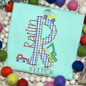 Może przedstawiać: Jasno błękitny t-shirt z literą R w kratę i imieniem "Ralin" wyszytym na czerwono. Litera R jest ozdobiona żółtym samochodem wywrotką, zielonym koparką i czerwonym samochodem budowlanym.