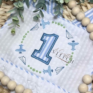May include: A white onesie with blue and white stripes, featuring a blue and white striped number one surrounded by a circle of blue airplanes and paper airplanes. The text "A Crafty Stitch" is embroidered in the bottom right corner.