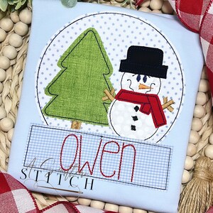 May include: A light blue t-shirt featuring a snowman and Christmas tree design. The snowman wears a black hat and red scarf. The name "Owen" is embroidered in red on a blue gingham patch. The shirt has a round design with a dotted background.