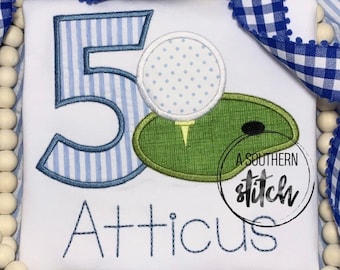 May include: White t-shirt featuring a blue striped number 5, a golf ball, and a green golf course design. The name "Atticus" is embroidered below the number. The shirt is decorated with blue and white gingham ribbon and wooden beads.