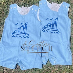 Peut inclure: Deux barboteuses bleues claires pour bébés avec un motif de bateau et des initiales brodées sur le devant. Les barboteuses ont des bordures blanches et des fermetures à boutons. Le texte "A Southern Stitch II" est visible.