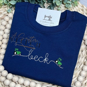 May include: A navy blue t-shirt with a white embroidered design that says "A Southern Stitch" and features a tractor and a needle and thread. The design also includes the word "beck" in a cursive font.