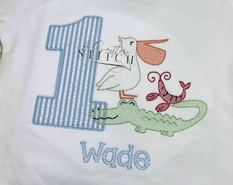 May include: A white onesie with blue and white striped number one, a pelican, alligator, and shrimp embroidered on it. The name Wade is embroidered below the animals.