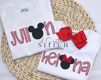 Puede incluir: Dos camisetas blancas con letras de vichy rojas y una cabeza de Mickey Mouse en blanco y negro. Las camisetas dicen "Julian" y "Helena".