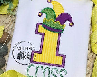 Puede incluir: Un pelele blanco con un número uno amarillo con contorno morado. El número uno tiene un sombrero de bufón verde y morado en la parte superior. La palabra "cross" está bordada en verde debajo del número.