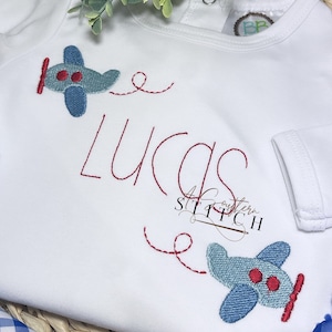 Pode incluir: Macacão branco para bebê com aviões bordados em vermelho e o nome "Lucas" em fio vermelho.