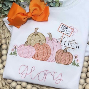 Könnte beinhalten: Weißes T-Shirt mit einem gestickten Design aus Kürbissen in Rosa-, Pfirsich- und Goldtönen. Das Shirt hat den Text "Glory" und ein "Zu verkaufen"-Schild. Oben befindet sich eine orangefarbene Schleife.