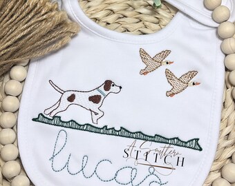 Op de afbeelding: Witte slabber met een geborduurd ontwerp van een hond die door gras rent met twee eenden die erboven vliegen. De naam "Lucas" is in cursief onder het ontwerp geborduurd. De tekst "A Southern Stitch" is onder de naam geborduurd.