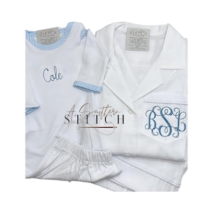 May include: White pyjama set with blue trim and embroidery. The set includes a long-sleeved top with a pocket and monogram, a short-sleeved onesie with the name "Cole", and matching shorts. The brand "A Southern Stitch" is visible.