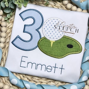 Puede incluir: Una camisa blanca bordada con un número 3 azul, un campo de golf verde y una bola de golf blanca con lunares azules. El texto "Emmett" está bordado debajo del diseño.