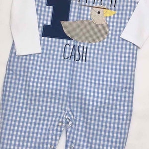May include: A blue and white gingham baby outfit with a white long-sleeved shirt and blue overalls. The overalls have a blue number one with a brown duck applique and the name "CASH" embroidered on the front.