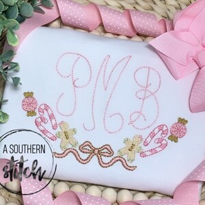 Puede incluir: Un pelele blanco con letras bordadas en rosa y un diseño de bastones de caramelo, galletas de jengibre y lazos. El pelele tiene el texto "A Southern Stitch" en la esquina inferior izquierda.