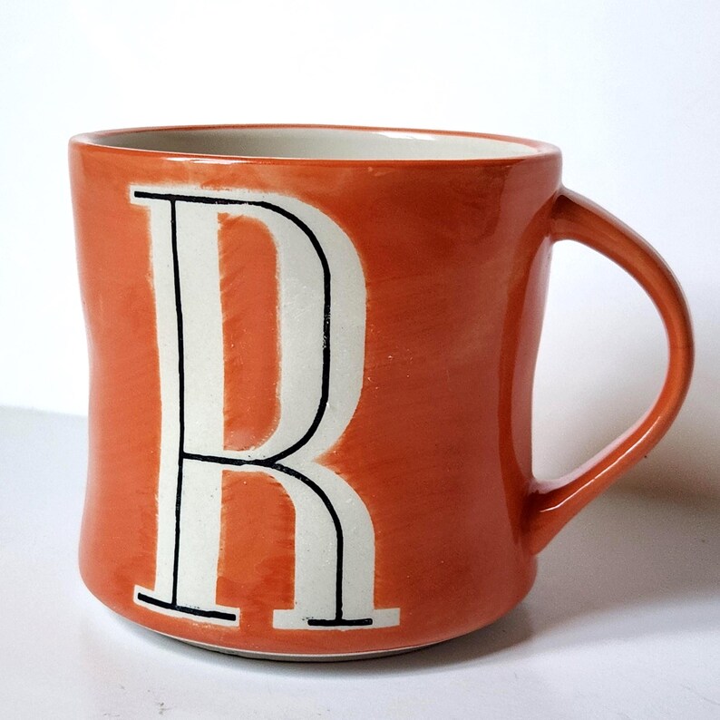 May include: Orange ceramic mug with a white letter "R" outlined in black. The mug has a curved handle and a white interior. The exterior is a solid orange color.