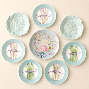 May include: A collection of ceramic plates including two light green leaf-shaped plates, four plates featuring illustrations of asparagus bundles tied with purple ribbons, and one central plate decorated with a colorful floral bouquet arrangement.