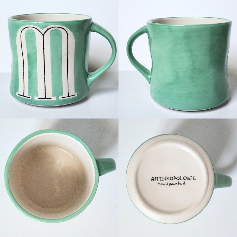 May include: A set of four ceramic mugs. One mug is green with a white "M" design. The other mugs are solid green. The interior of the mugs is cream-colored. The bottom of one mug has the text "ANTHROPOLOGIE hand painted".