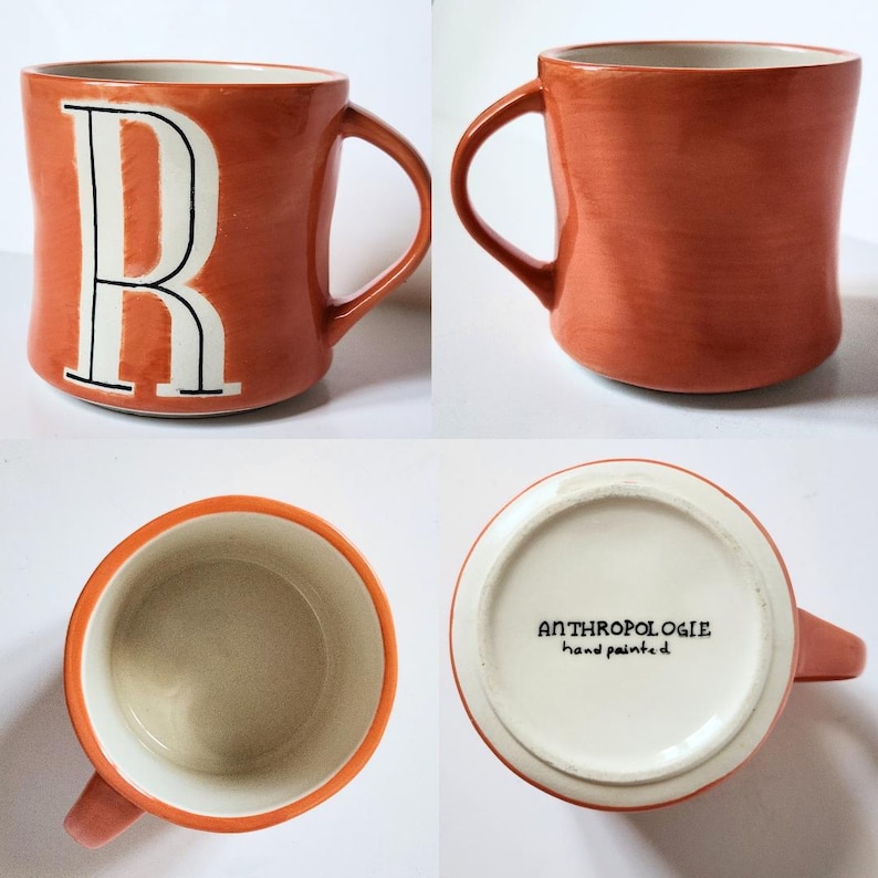 May include: A terracotta-colored ceramic mug with a white letter "R" outlined in black. The mug has a curved handle and a cream-colored interior. The bottom of the mug reads "ANTHROPOLOGIE hand painted".