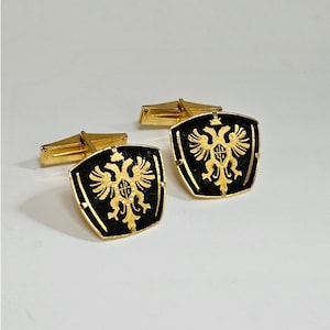 Puede incluir: Un par de gemelos dorados con un diseño en forma de escudo sobre un fondo negro. Cada gemelo muestra un emblema dorado de un águila bicéfala centrado en el escudo, con un cierre de palanca de metal dorado para una sujeción segura.