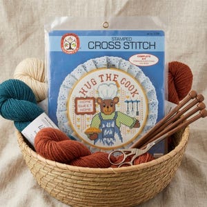 May include: A cross-stitch kit with a bear chef design, reading "Hug the Cook," includes yarn, needles, and scissors. The kit is packaged in a clear plastic bag and presented in a woven basket. The kit includes everything you need to complete the project.