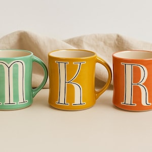 May include: Three ceramic mugs in a row, each with a different color and letter. The first mug is mint green with the letter "M", the second is yellow with a "K", and the third is orange with an "R". Each letter is white with a black outline.