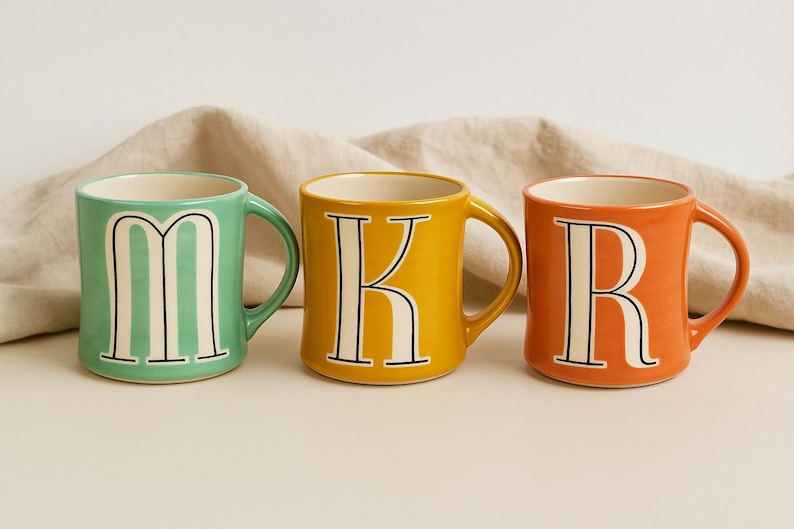 May include: Three ceramic mugs in a row, each with a different color and letter. The first mug is mint green with the letter "M", the second is yellow with a "K", and the third is orange with an "R". Each letter is white with a black outline.