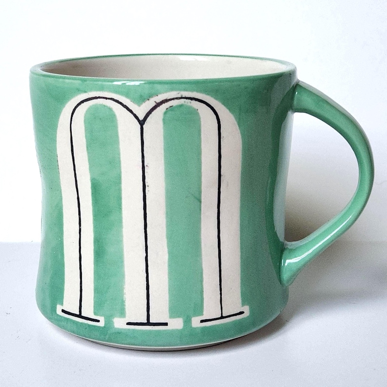 May include: A green ceramic mug with a white letter "M" design. The letter is outlined in black. The mug has a curved handle and a white interior.