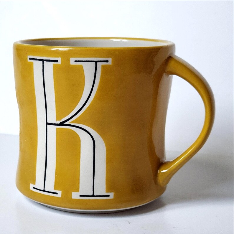 May include: Mustard yellow ceramic mug with a white letter "K" in a bold, serif font. The mug has a curved handle and a white interior. The mug is a standard size for coffee or tea.