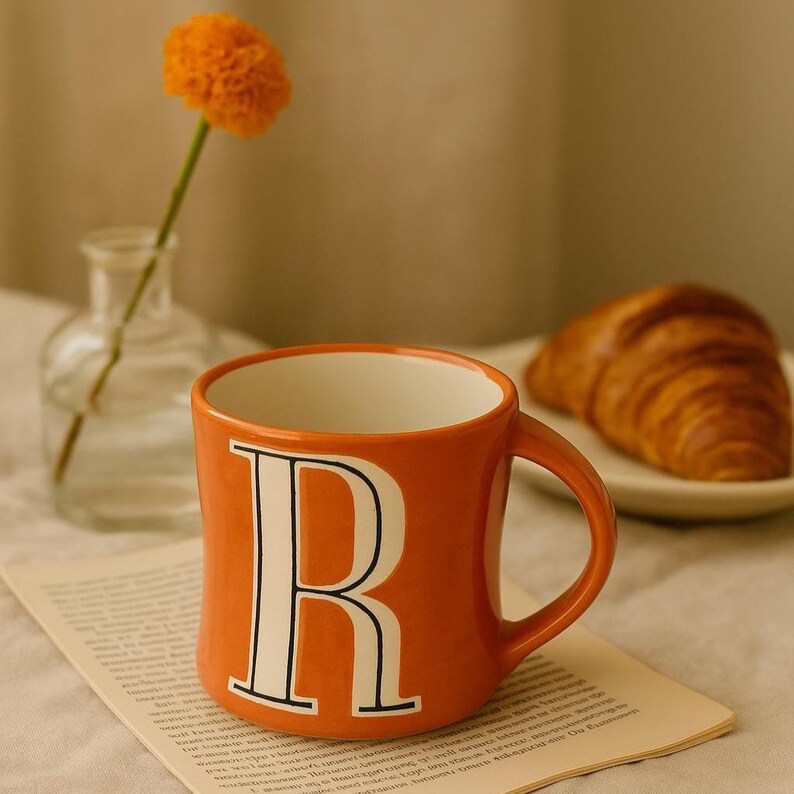 May include: Orange ceramic mug with a white interior and a large white letter "R" on the front. A croissant sits on a white plate in the background, with a small vase and flower to the left.
