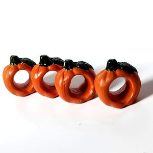 May include: A set of four orange pumpkin-shaped napkin rings, each featuring a dark green stem and leaf detail on top. The rings have a smooth, rounded exterior and a circular opening in the center for holding napkins securely in place.