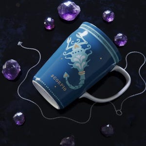 K&ouml;nnte beinhalten: Eine blaue Keramiktasse mit einem Skorpion-Sternzeichen-Design und einem wei&szlig;en Henkel. Die Tasse ist von lila Kristallen und einer silbernen Kette umgeben. Das Wort "SCORPIO" ist auf die Tasse gedruckt.