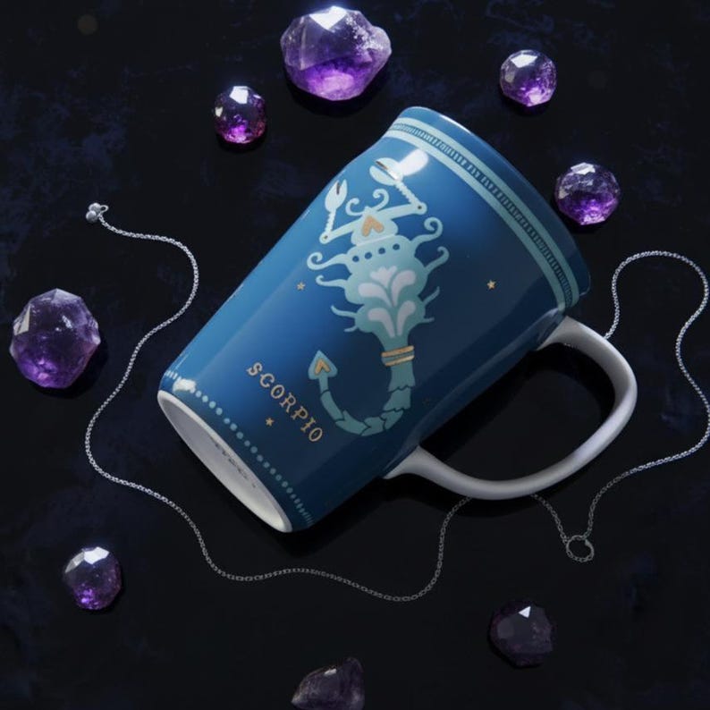 K&ouml;nnte beinhalten: Eine blaue Keramiktasse mit einem Skorpion-Sternzeichen-Design und einem wei&szlig;en Henkel. Die Tasse ist von lila Kristallen und einer silbernen Kette umgeben. Das Wort "SCORPIO" ist auf die Tasse gedruckt.