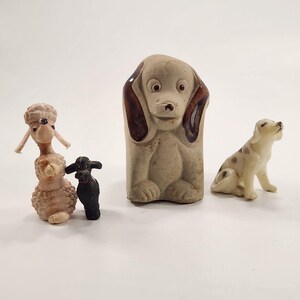 May include: Four dog figurines in various styles. A light brown poodle, a small black dog, a large beige dog with brown ears, and a small white dog with brown spots. All are standing on a white surface.