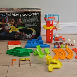 Puede incluir: Un set de juguetes Playrail by Tomy Merry-Go-Copter que incluye una caja, una torre alta naranja con un helicóptero, una locomotora roja y blanca, un vagón blanco, cuatro figuras pequeñas, vías curvas naranjas y un letrero.