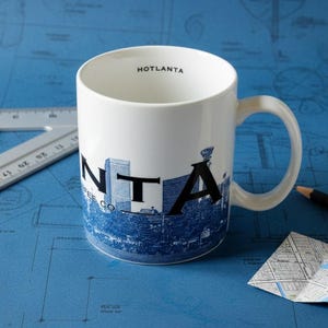 Könnte beinhalten: Weiße Keramik-Kaffeetasse mit blauem und schwarzem Skyline-Design. Die Tasse trägt den Schriftzug "HOTLANTA" am Rand und "NTA COFFEE CO" an der Seite. Die Tasse steht auf einer blauen Oberfläche.