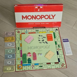 May include: A Monopoly property trading board game set including a red and white box, a game board with green and white squares, various denominations of paper money, metal tokens, red and green plastic houses and hotels, dice, and cards.