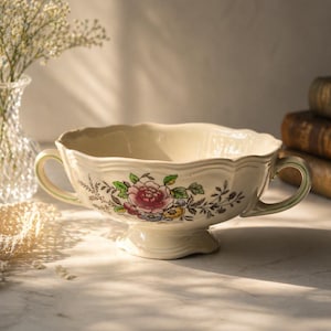 May include: Cream-coloured ceramic serving bowl with a scalloped rim and two handles. The bowl features a floral design with pink, red, and green accents. The bowl sits on a pedestal base, and is likely vintage.