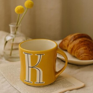 May include: A yellow ceramic mug with a white interior and a large white letter "K" on the front. The mug is on a piece of paper, with a croissant and a small vase with yellow flowers in the background.
