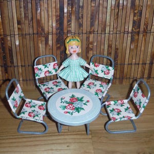 May include: A vintage doll stands near a miniature patio set. The set includes a round table and four chairs with floral print fabric. The doll wears a green dress. The background is a bamboo blind.