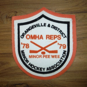 May include: Orangeville & District Minor Hockey Association patch with a white background, black border, and orange stitching. The patch features the text "OMHA REPS '78 '79" and "MINOR PEE WEE ASSOCIATION" along with a pair of crossed hockey sticks.