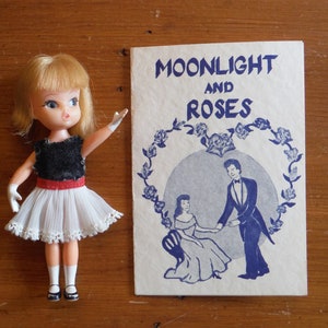 May include: A vintage doll wearing a white tulle skirt with a black bodice and red belt, holding a white glove. The doll is standing next to a vintage book with the title "Moonlight and Roses" printed in blue.